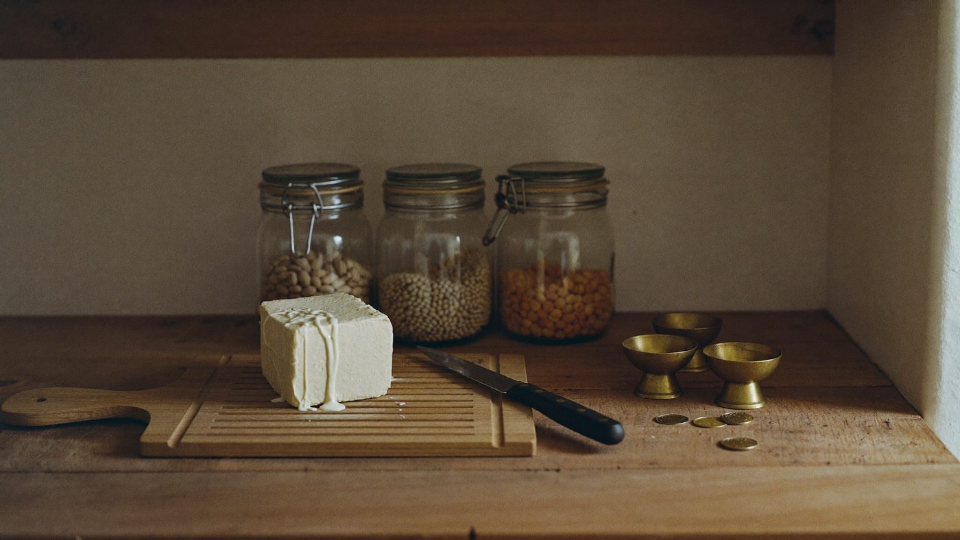 Un blocco di tofu su un tagliere, dietro barattoli di legumi e monete.